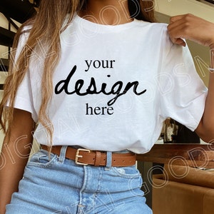 Op de afbeelding: Een wit T-shirt met de tekst "your design here" gedrukt op de voorkant. Het shirt wordt gedragen door een persoon met lang bruin haar en wordt gecombineerd met een blauwe jeans en een bruine riem.