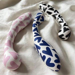 Könnte beinhalten: Drei gebogene Sexspielzeuge im Vibrator-Stil. Eines ist weiß mit rosa Herzen, ein anderes ist weiß mit blauen Herzen und das dritte ist weiß mit schwarzen Herzen. Sie sind auf einer weißen, strukturierten Oberfläche angeordnet.