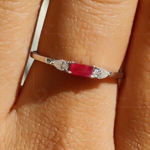 Könnte beinhalten: Ein zarter Silberring mit einem rechteckigen, leuchtend roten Edelstein, flankiert von zwei kleineren, birnenförmigen weißen Edelsteinen. Der Ring ist auf einem schmalen Band gefasst und zeigt ein minimalistisches Design.