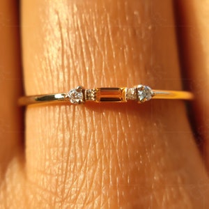 May include: A thin gold ring featuring a central rectangular baguette-cut orange gemstone flanked by two small round clear stones. The band is smooth and metallic, showcasing a minimalist design with three distinct stones set horizontally.