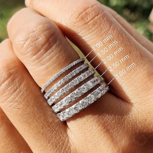 Puede incluir: Cinco anillos de eternidad plateados con piedras transparentes, apilados juntos. Los anillos están etiquetados con anchos de 1,00 mm, 1,50 mm, 2,00 mm, 2,50 mm y 3,00 mm, indicando el grosor variable de cada anillo individual del conjunto.