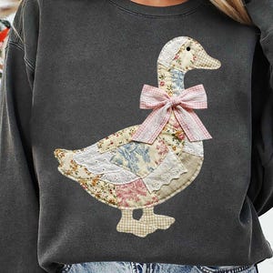 Peut inclure: Sweat-shirt gris foncé avec un motif d'oie en patchwork. L'oie est faite de tissus à motifs floraux et à motifs, avec un nœud rose à carreaux. Le motif comprend de la dentelle et un bec et des pieds à carreaux. Un haut confortable et décontracté.