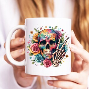 Peut inclure: Une tasse en c&eacute;ramique blanche avec un motif de cr&acirc;ne de sucre color&eacute;. Le cr&acirc;ne est d&eacute;cor&eacute; de fleurs et a un geste de main de signe de paix.
