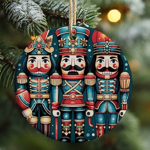 May include: A round Christmas ornament featuring three detailed nutcracker figures in teal, red, and gold. The ornament has a beige string for hanging and is set against a blurred background of a Christmas tree.