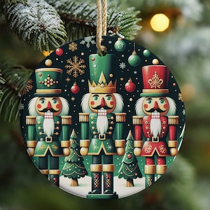 May include: A round Christmas ornament featuring three nutcracker soldiers in green and red uniforms. The ornament has a dark green background with snowflakes and Christmas tree accents. A beige string is attached for hanging.