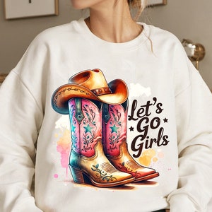Op de afbeelding: Witte sweatshirt met een aquarelontwerp van roze en bruine cowboylaarzen en een bruine cowboyhoed. De tekst "Let's Go Girls" is in het zwart gedrukt. Een grafische afbeelding met countrythema.
