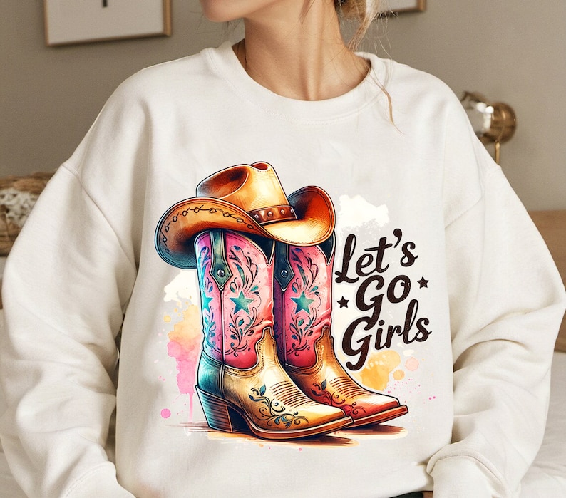 Op de afbeelding: Witte sweatshirt met een aquarelontwerp van roze en bruine cowboylaarzen en een bruine cowboyhoed. De tekst "Let's Go Girls" is in het zwart gedrukt. Een grafische afbeelding met countrythema.