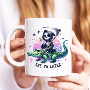 May include: White ceramic mug with a colorful graphic of a skeleton riding a green alligator. The skeleton is wearing a black hooded robe and holding a scythe. The text "See Ya Later" is printed below the graphic.