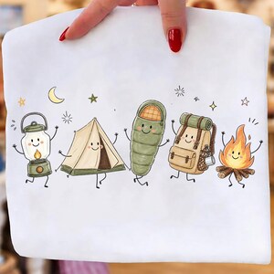 Puede incluir: Una camiseta blanca con un estampado gr&aacute;fico de cinco art&iacute;culos de camping antropom&oacute;rficos: una linterna, una tienda de campa&ntilde;a, un saco de dormir, una mochila y una hoguera. Cada objeto tiene una cara sonriente, extremidades finas y estrellas.