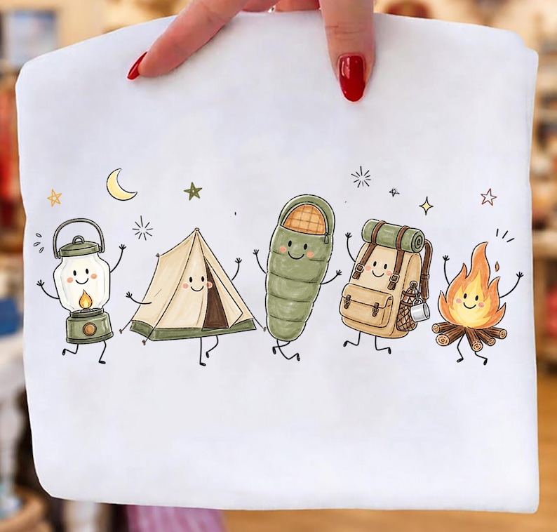 Puede incluir: Una camiseta blanca con un estampado gr&aacute;fico de cinco art&iacute;culos de camping antropom&oacute;rficos: una linterna, una tienda de campa&ntilde;a, un saco de dormir, una mochila y una hoguera. Cada objeto tiene una cara sonriente, extremidades finas y estrellas.