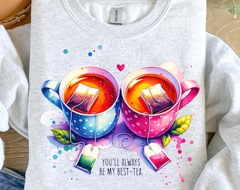 May include: A light grey sweatshirt with a watercolour design of two teacups with teabags. One cup is blue with white polka dots, the other is pink with white polka dots. The text "YOU'LL ALWAYS BE MY BEST-TEA" is below the cups.