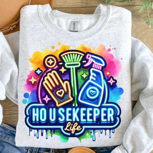 Op de afbeelding: Een grijs sweatshirt met een kleurrijk grafisch ontwerp van een reinigingsfles, een bezem en een schoonmaakhandschoen. De grafiek is in neon-stijl met de tekst "HOUSEKEEPER Life".