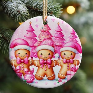May include: Round Christmas ornament featuring three gingerbread people wearing pink Santa hats and bow ties, with pink Christmas trees in the background. The ornament is decorated with snowflakes and a pink gift.