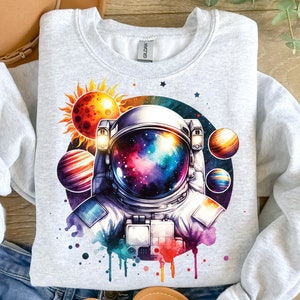 Może przedstawiać: Aquarela przedstawiająca astronautę z galaktyką w hełmie. Astronautę otaczają planety i słońce. Ilustracja znajduje się na szarej bluzie.