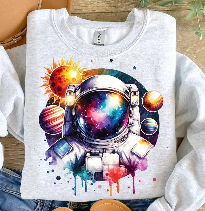 Może przedstawiać: Aquarela przedstawiająca astronautę z galaktyką w hełmie. Astronautę otaczają planety i słońce. Ilustracja znajduje się na szarej bluzie.