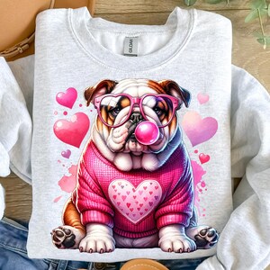 K&ouml;nnte beinhalten: Ein grauer Pullover mit einem rosa und wei&szlig;en Bulldoggen, der eine rosa herzf&ouml;rmige Sonnenbrille tr&auml;gt und eine rosa Blase bl&auml;st. Der Bulldogge tr&auml;gt einen rosa Pullover mit einem rosa Herzen auf der Vorderseite. Der Hintergrund ist wei&szlig; mit rosa Herzen.