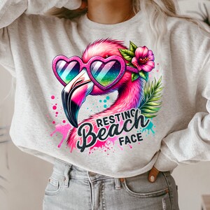 May include: A grey sweatshirt featuring a pink flamingo wearing heart-shaped sunglasses and a pink flower. The flamingo is surrounded by colourful paint splatters and the text "Resting Beach Face".