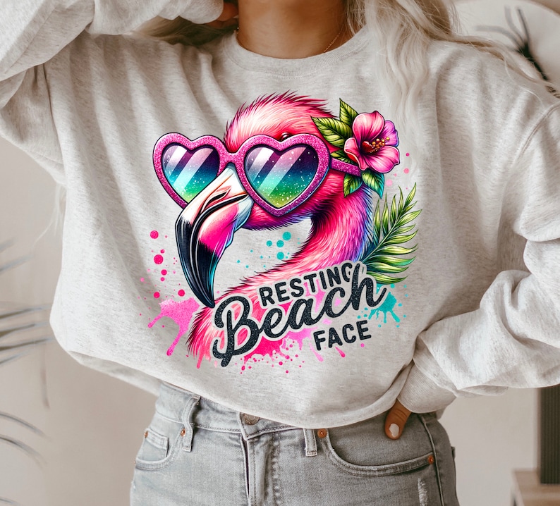May include: A grey sweatshirt featuring a pink flamingo wearing heart-shaped sunglasses and a pink flower. The flamingo is surrounded by colourful paint splatters and the text "Resting Beach Face".