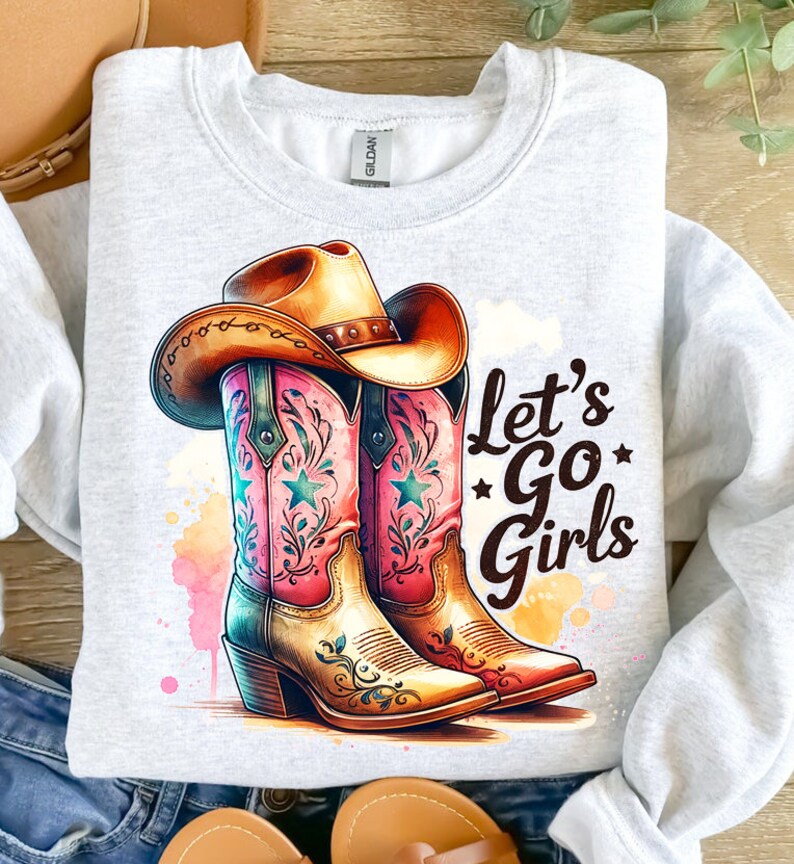 Op de afbeelding: Een lichtgrijs sweatshirt met een aquarelontwerp van een cowboyhoed en -laarzen. De laarzen zijn roze en turquoise met ster- en bloemaccenten. De tekst "Let's Go Girls" is in het zwart gedrukt.