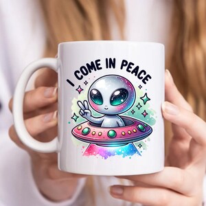 May include: White ceramic mug with a colorful illustration of a cartoon alien in a flying saucer. The alien is waving with one hand and the text "I come in peace" is written above the saucer.