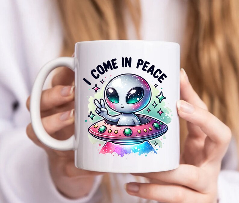 May include: White ceramic mug with a colorful illustration of a cartoon alien in a flying saucer. The alien is waving with one hand and the text "I come in peace" is written above the saucer.