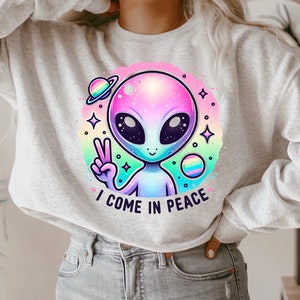 Puede incluir: Una sudadera gris claro con un gr&aacute;fico de extraterrestre colorido y el texto "Vengo en paz".