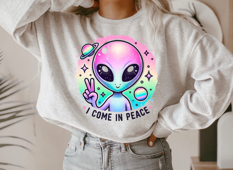 Puede incluir: Una sudadera gris claro con un gr&aacute;fico de extraterrestre colorido y el texto "Vengo en paz".
