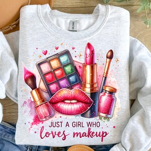 May include: A gray sweatshirt with a colorful graphic design of makeup items, including a lipstick, eyeshadow palette, and makeup brushes. The text "Just a girl who loves makeup" is printed below the graphic.