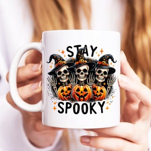 Puede incluir: Taza de cer&aacute;mica blanca con un dise&ntilde;o de Halloween. El dise&ntilde;o presenta tres figuras de esqueleto con sombreros de bruja, sosteniendo calabazas y las palabras "STAY SPOOKY". El dise&ntilde;o es en naranja, negro y blanco.