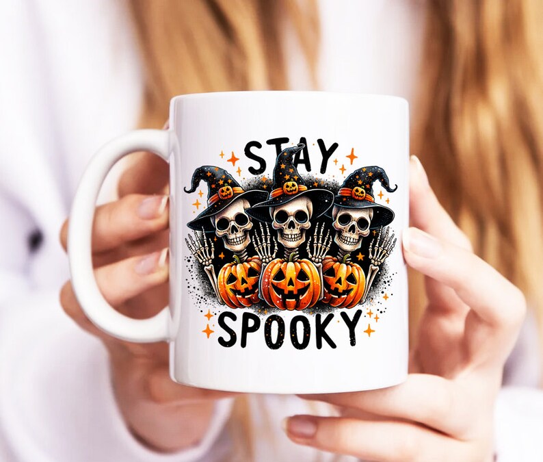 Puede incluir: Taza de cer&aacute;mica blanca con un dise&ntilde;o de Halloween. El dise&ntilde;o presenta tres figuras de esqueleto con sombreros de bruja, sosteniendo calabazas y las palabras "STAY SPOOKY". El dise&ntilde;o es en naranja, negro y blanco.