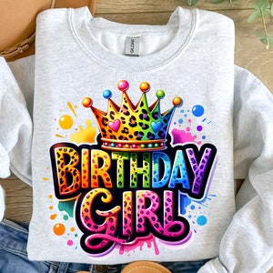 May include: A gray sweatshirt with a colorful graphic design of a crown, the words "Birthday Girl" and a leopard print pattern. The design is surrounded by colorful paint splatters.