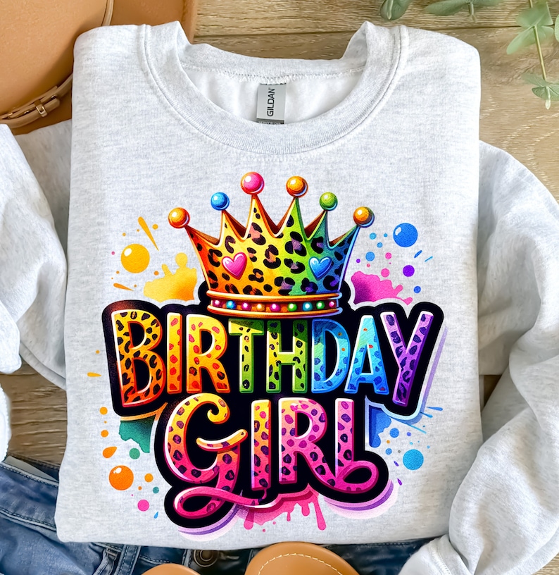 May include: A gray sweatshirt with a colorful graphic design of a crown, the words "Birthday Girl" and a leopard print pattern. The design is surrounded by colorful paint splatters.
