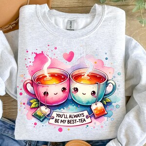 May include: A light gray sweatshirt with a watercolor design of two teacups with smiling faces. The teacups are holding hands and have steam coming out of them. The text "You'll always be my best-tea" is written below the teacups.