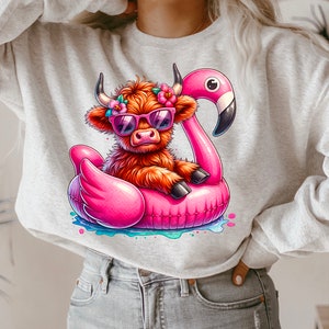 May include: A gray sweatshirt with a cartoon illustration of a Highland cow wearing sunglasses and riding a pink flamingo pool float. The cow has a flower crown on its head.