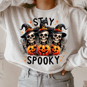 Puede incluir: Una sudadera gris claro con un dise&ntilde;o de Halloween. El gr&aacute;fico presenta tres cabezas de esqueleto con sombreros de bruja, sosteniendo calabazas naranjas y las palabras "STAY SPOOKY".