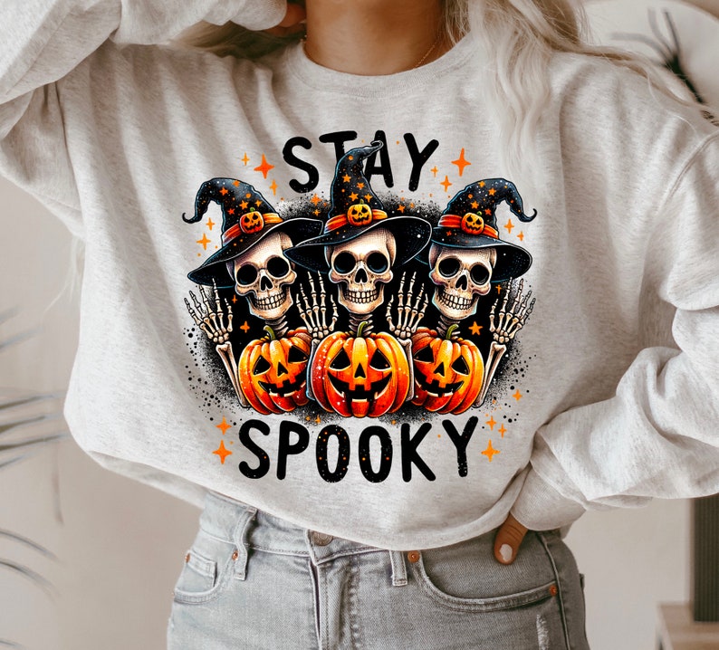 Puede incluir: Una sudadera gris claro con un dise&ntilde;o de Halloween. El gr&aacute;fico presenta tres cabezas de esqueleto con sombreros de bruja, sosteniendo calabazas naranjas y las palabras "STAY SPOOKY".
