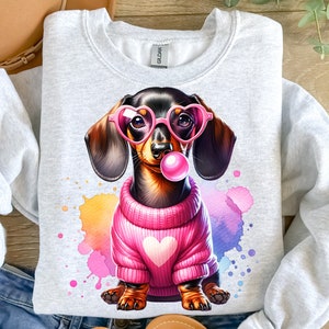 May include: A gray sweatshirt with a cartoon image of a dachshund wearing pink heart-shaped sunglasses and a pink sweater with a heart on it. The dachshund is blowing a pink bubble. The background is a watercolor design with pink, yellow, and blue colors.