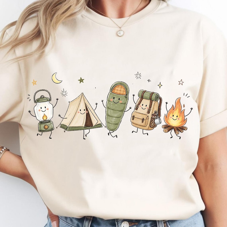 Puede incluir: Una camiseta de color crema con un estampado gr&aacute;fico de cinco art&iacute;culos de camping antropom&oacute;rficos: una linterna, una tienda de campa&ntilde;a, un saco de dormir, una mochila y una hoguera, todos con caras sonrientes, rodeados de estrellas y una luna creciente.