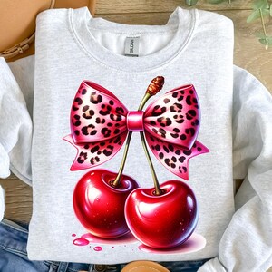 May include: A light gray sweatshirt featuring a vibrant illustration of two glossy red cherries with a pink bow. The bow has a leopard print design. The cherries have green stems and are set against a white background.