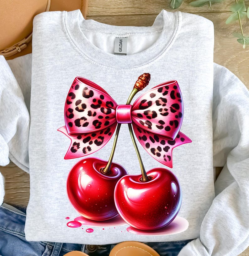 May include: A light gray sweatshirt featuring a vibrant illustration of two glossy red cherries with a pink bow. The bow has a leopard print design. The cherries have green stems and are set against a white background.