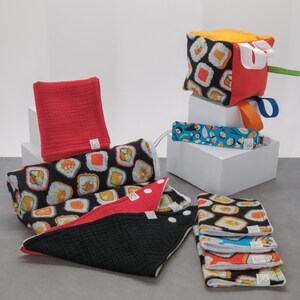 Op de afbeelding: Een verzameling babyaccessoires, waaronder slabbetjes, een kubus speelgoed en spuugdoekjes. De items hebben sushi roll patronen in zwart, wit, oranje en groen. Een rode vierkante doek en een blauwe fopspeenclip zijn ook aanwezig.