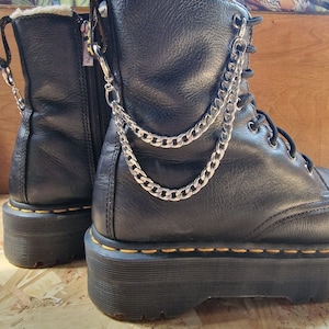 May include: Black leather platform boots with a silver chain attached to the top of each boot. The boots have a yellow stitch around the platform sole.