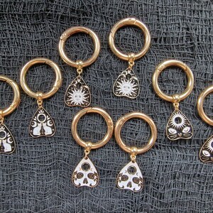 May include: Eight gold-toned keyrings with black and white enamel charms. The charms are shaped like a triangle and feature different designs, including a sun, a skull, and cats.