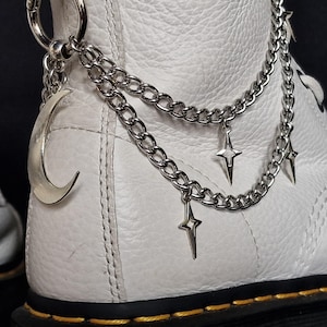 May include: Close-up of a white leather boot with silver chain accessories. The chains feature a crescent moon charm and star pendants. The boot has black soles and stitching.