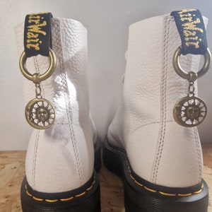 May include: White leather boots with black laces and a black platform sole. The boots have a gold charm with a sun and moon design attached to a black strap.