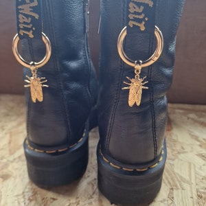 May include: Black leather boots with gold accents and a gold insect charm attached to a gold ring. The boots have a yellow and black "Dr. Martens" tag on the back.