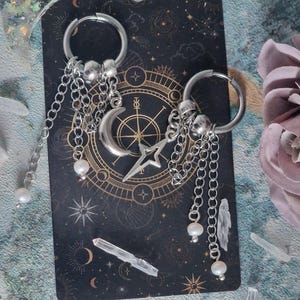 May include: Silver hoop earrings with hanging chains, faux pearls, and celestial charms. Each earring has a crescent moon, a star, and a compass rose design. The earrings are shown on a dark background with a celestial theme.