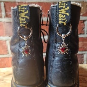 May include: A pair of black leather boots with a red heart charm attached to each boot. The charms are silver with a red heart in the centre and a silver starburst around the heart. The boots have a black fur lining and a yellow strap with the text "Dr. Martens" on it.