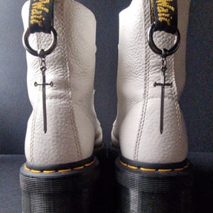 May include: White leather boots with black soles and yellow stitching. A silver sword charm hangs from a black strap on each boot.