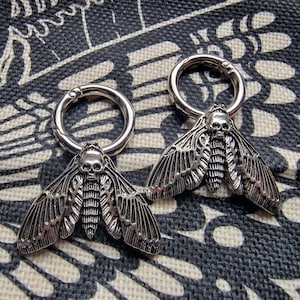 May include: Two silver skull moth earrings with a circular hoop design. The moths have detailed wings and a skull-shaped head.
